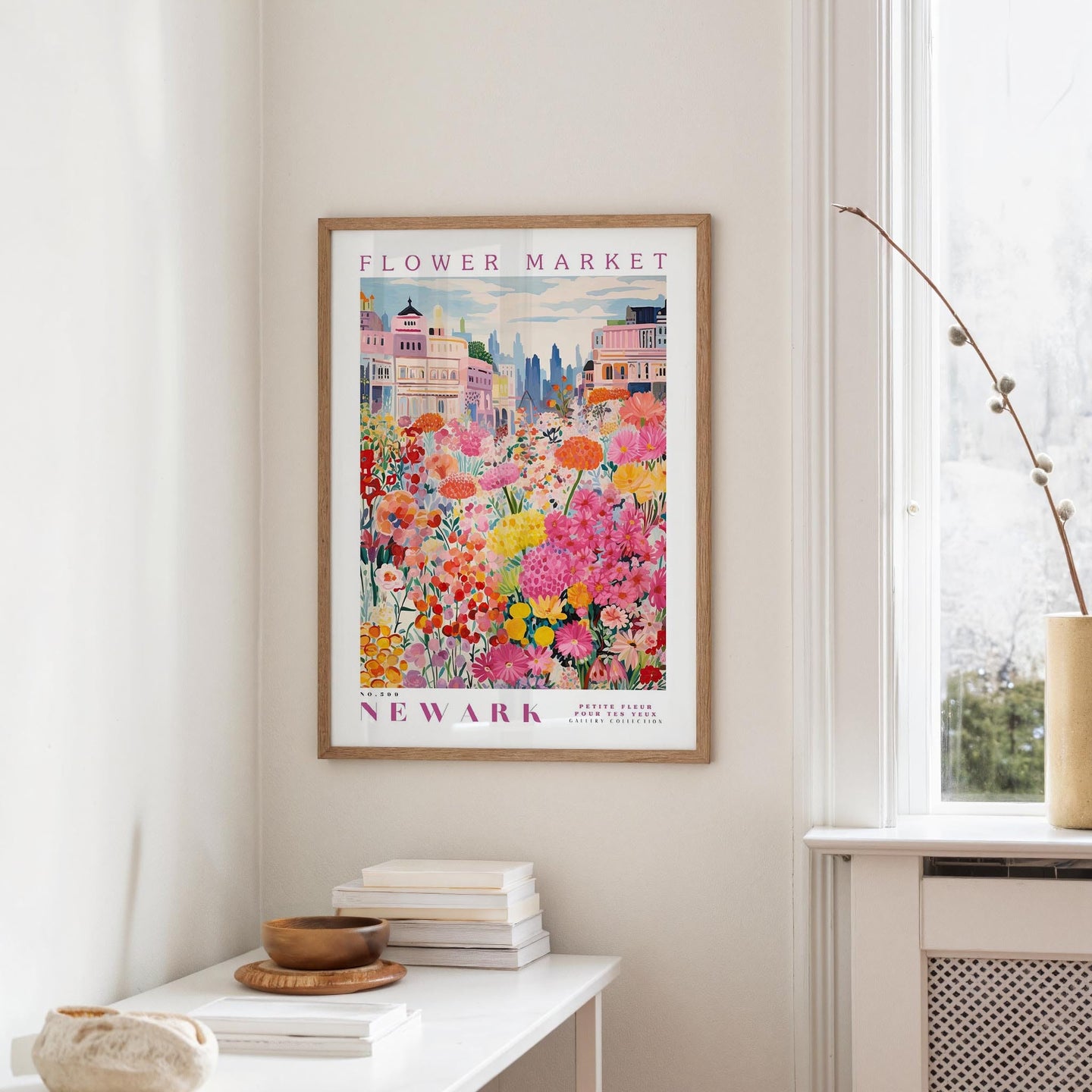 a framed poster of a colorful floral scene hanging on a wall, with a white table and a vase of flowers in the foreground.