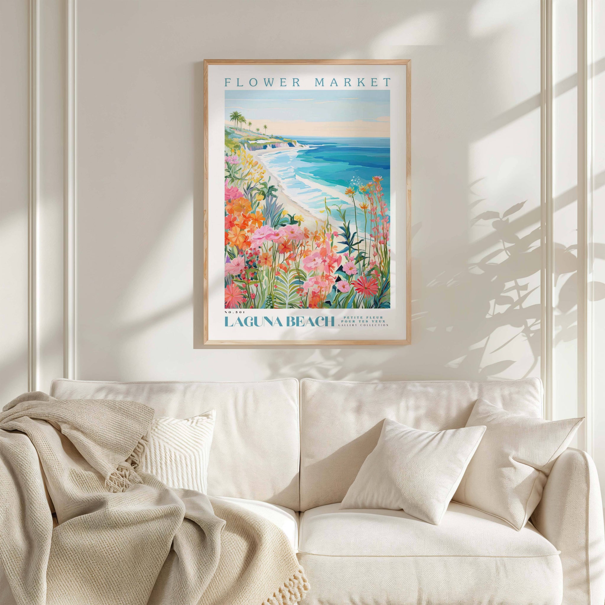 a framed poster of a beach scene with flowers hanging on a wall above a white couch with pillows.