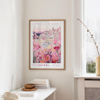 a framed poster of a cityscape with a flower market in the background, hanging on a wall in a room with a window and a small table with books and a bowl on it.