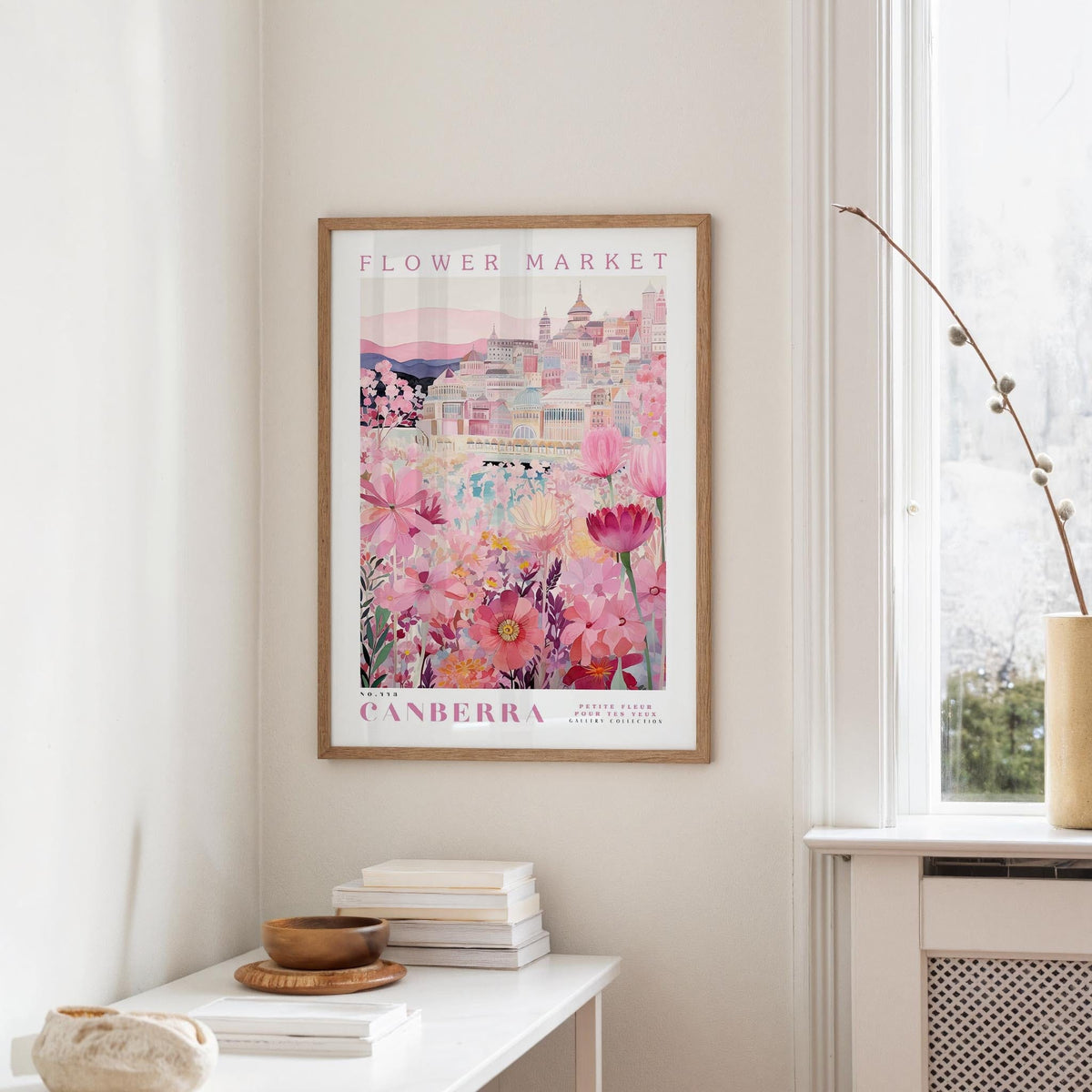 a framed poster of a cityscape with a flower market in the background, hanging on a wall in a room with a window and a small table with books and a bowl on it.