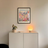 a white cabinet with a potted plant and a lamp on top, set against a wall with a framed poster of a flower market in Buffalo.