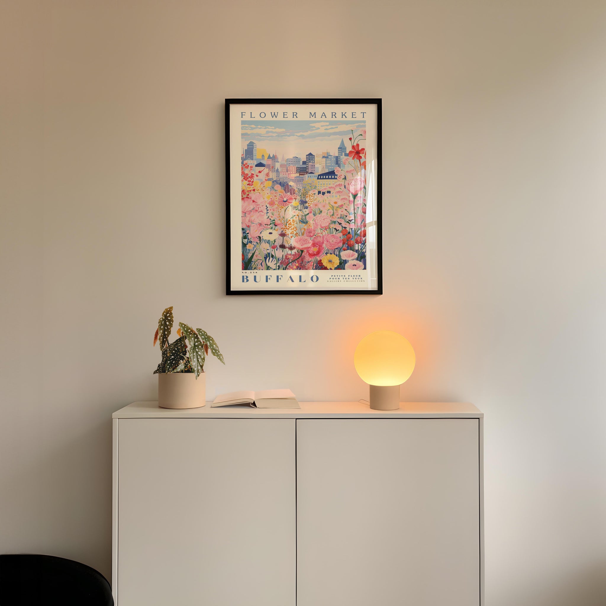 a white cabinet with a potted plant and a lamp on top, set against a wall with a framed poster of a flower market in Buffalo.