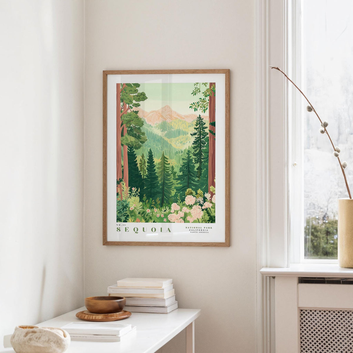 a framed poster of a lush forest scene with mountains in the background, hanging on a wall in a room with a white table and a window.