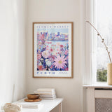 a framed poster of a cityscape with a floral design, hanging on a wall in a room with a window and a small table with books and a bowl on it.