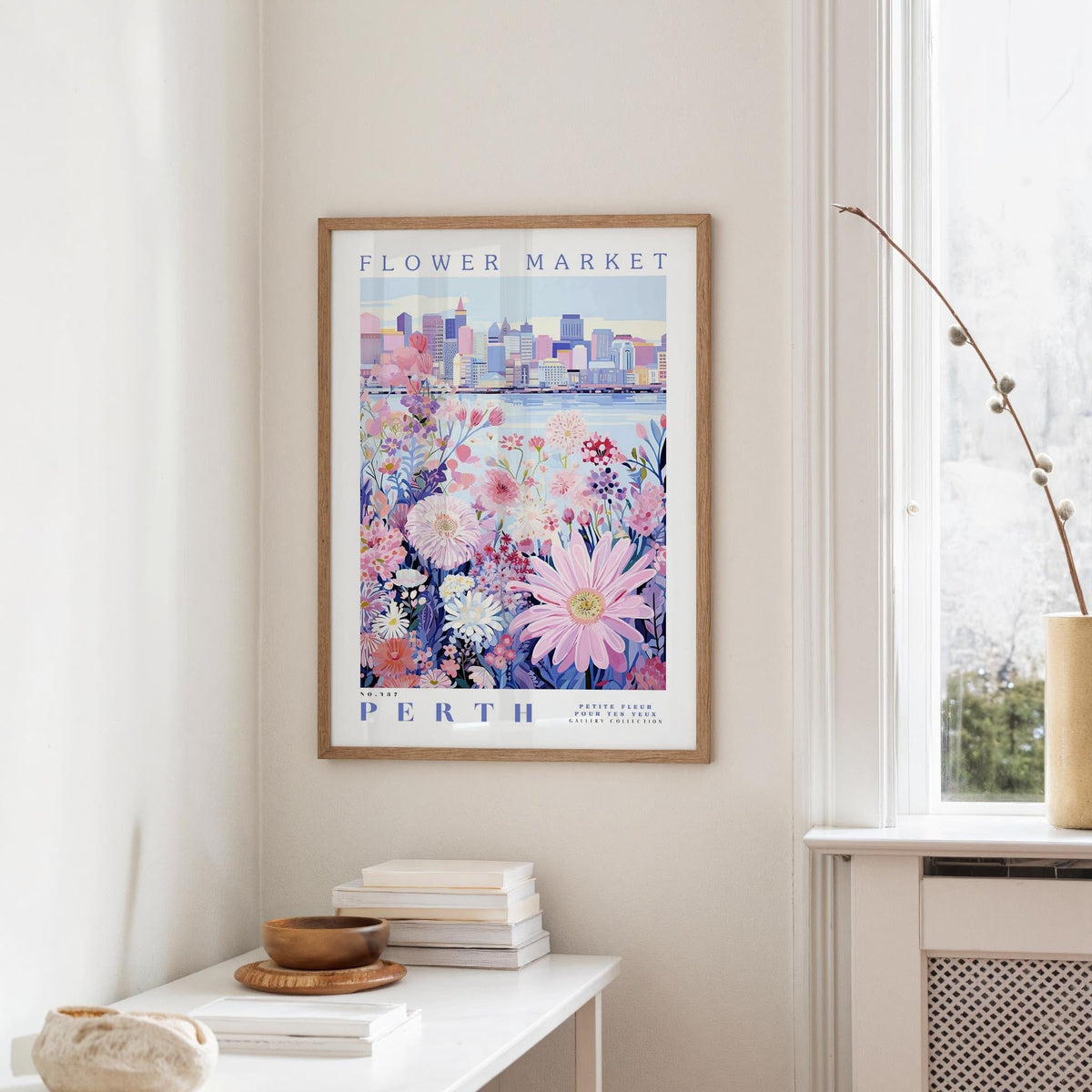 a framed poster of a cityscape with a floral design, hanging on a wall in a room with a window and a small table with books and a bowl on it.