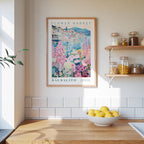 a kitchen scene with a framed poster of a flower market in Säalito hanging on the wall above a wooden counter. On the counter, there is a bowl of lemons and a plant. The kitchen has white tile walls and a wooden shelf with various jars and containers.