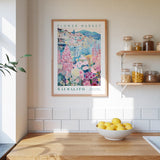 a kitchen scene with a framed poster of a flower market in Säalito hanging on the wall above a wooden counter. On the counter, there is a bowl of lemons and a plant. The kitchen has white tile walls and a wooden shelf with various jars and containers.