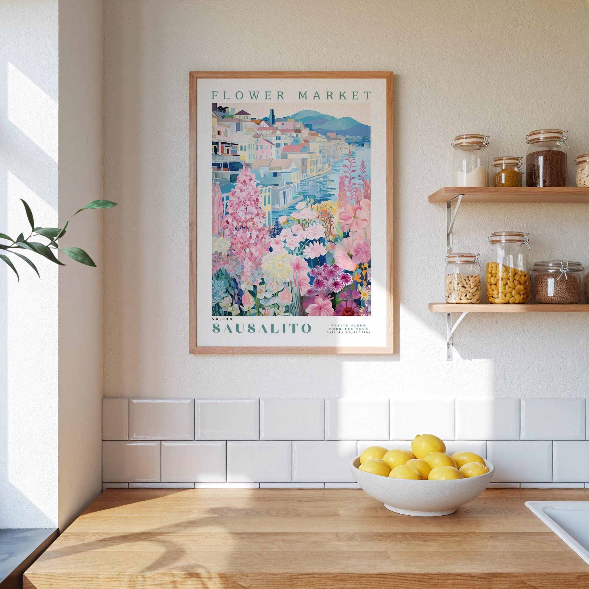 a kitchen scene with a framed poster of a flower market in Säalito hanging on the wall above a wooden counter. On the counter, there is a bowl of lemons and a plant. The kitchen has white tile walls and a wooden shelf with various jars and containers.