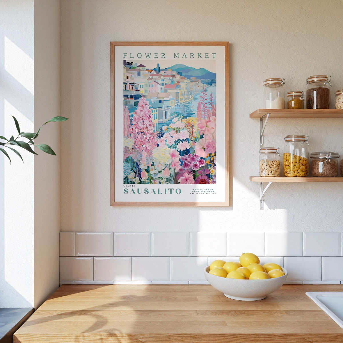 a kitchen scene with a framed poster of a flower market in Säalito hanging on the wall above a wooden counter. On the counter, there is a bowl of lemons and a plant. The kitchen has white tile walls and a wooden shelf with various jars and containers.