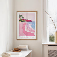 a framed poster of a beach scene with palm trees and a pink and blue ocean, hanging on a wall in a room with a white table and a plant.