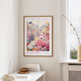 a framed poster of a colorful cityscape with flowers hanging on a wall in a room with a white table and a window.