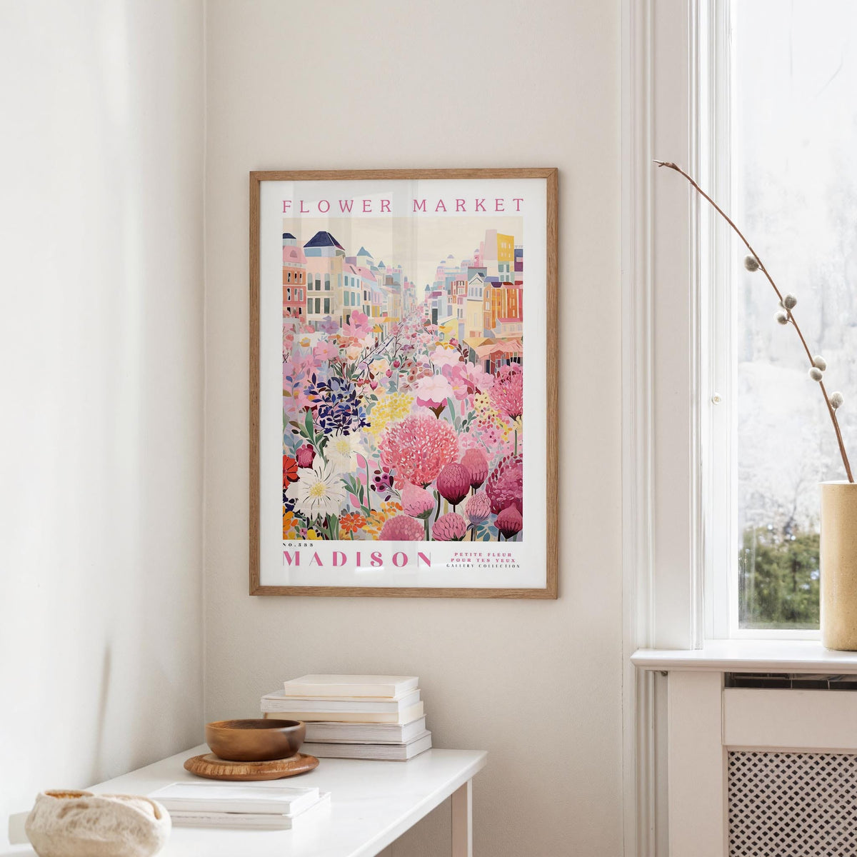 a framed poster of a colorful cityscape with flowers hanging on a wall in a room with a white table and a window.