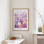 a framed poster of a colorful cityscape with flowers hanging on a wall, next to a window and a small table with books and a bowl.