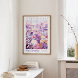 a framed poster of a colorful cityscape with flowers hanging on a wall, next to a window and a small table with books and a bowl.