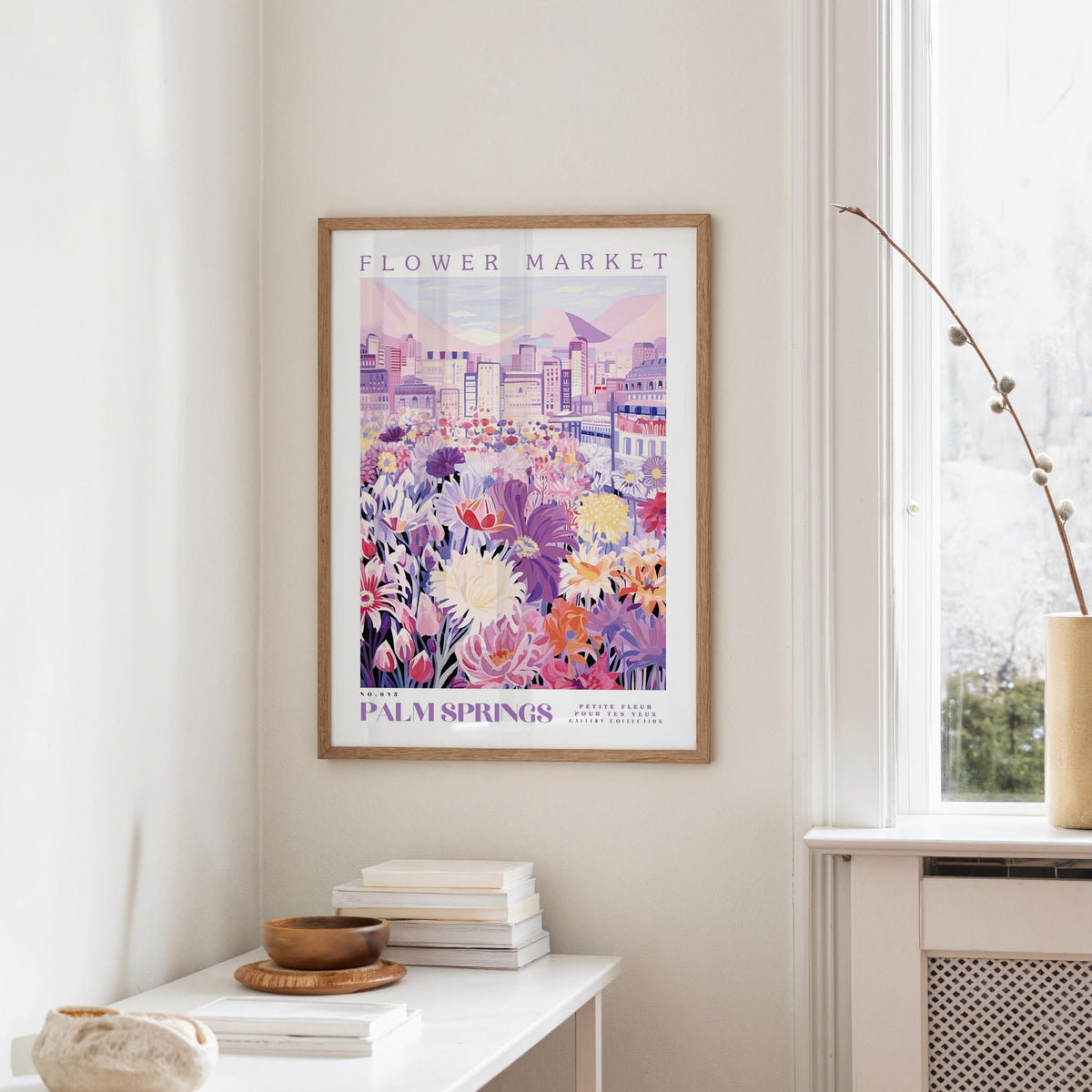 a framed poster of a colorful cityscape with flowers hanging on a wall, next to a window and a small table with books and a bowl.