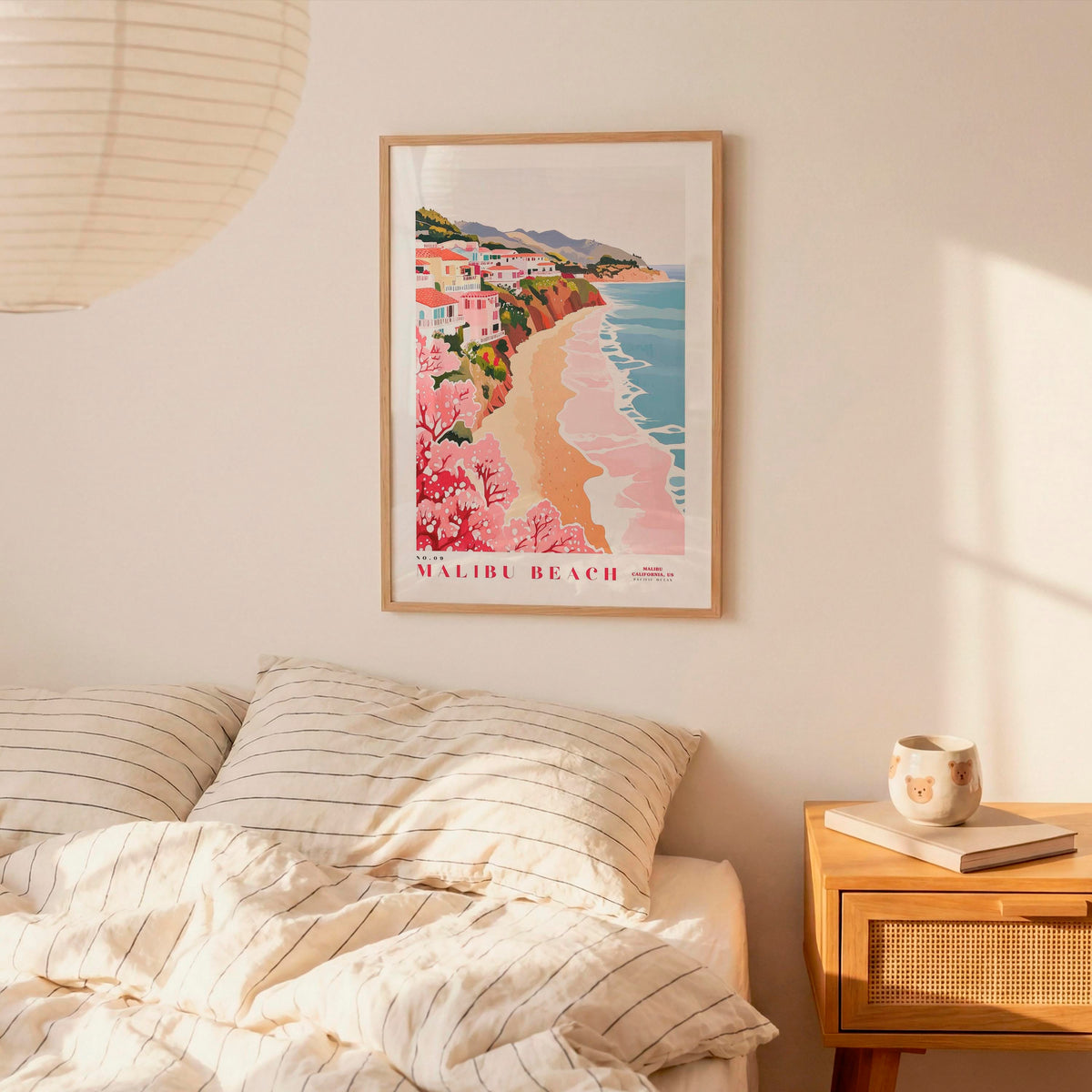 a framed poster of a beach scene with a coastal town, hanging on a wall above a bed with striped pillows.