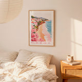 a framed poster of a beach scene with a coastal town, hanging on a wall above a bed with striped pillows.