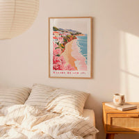 a framed poster of a beach scene with a coastal town, hanging on a wall above a bed with striped pillows.