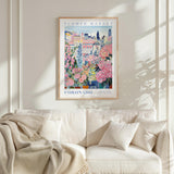 a framed poster of a colorful floral scene hanging on a wall above a white couch with a blanket draped over it.