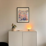 a white cabinet with a potted plant and a lamp on top, set against a white wall with a framed poster of a floral design hanging above it.