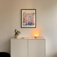 a white cabinet with a potted plant and a lamp on top, set against a white wall with a framed poster of a floral design hanging above it.
