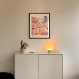 a white cabinet with a potted plant and a lamp on top, set against a wall with a framed poster of a cityscape above it.