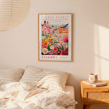 a bedroom setting with a framed poster of a colorful flower market hanging on the wall above a bed with striped pillows.