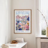 a framed poster of a colorful floral scene hanging on a wall, with a white table and a vase of flowers in the foreground.