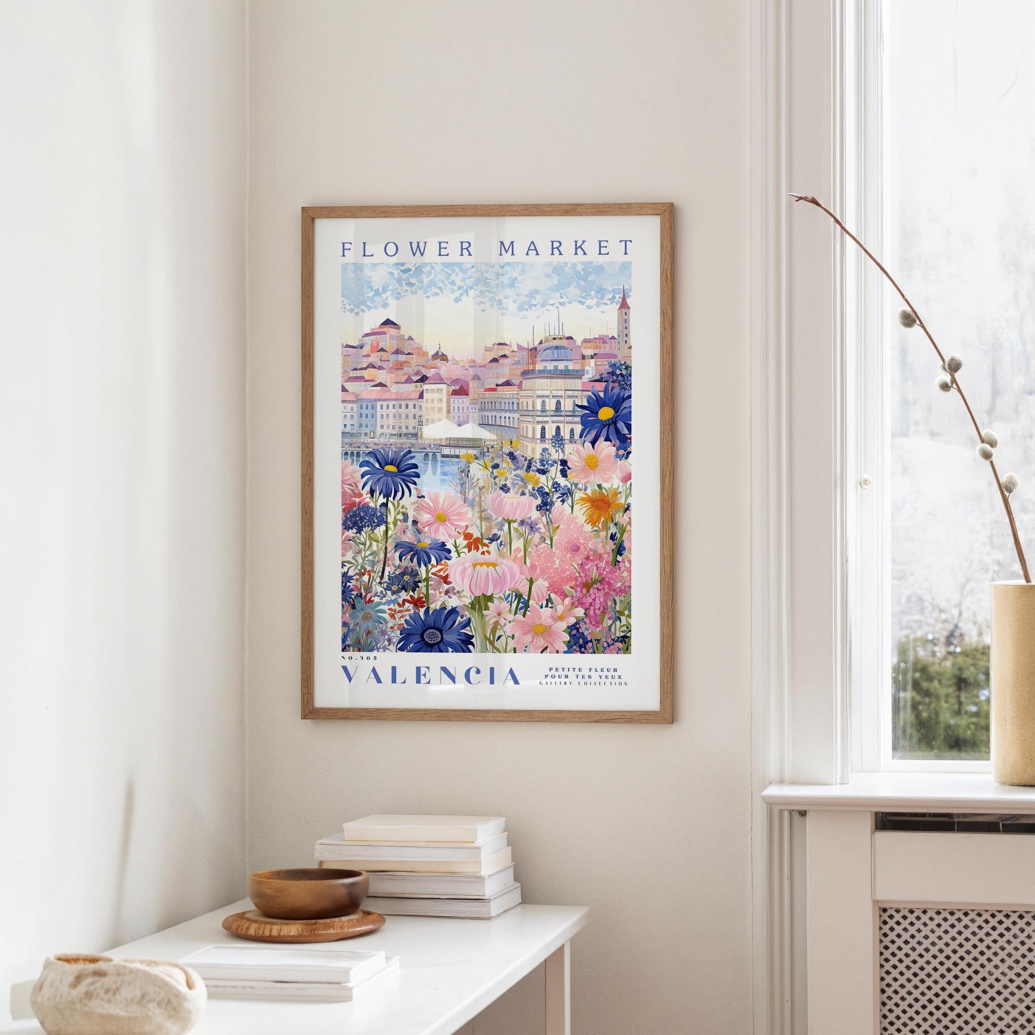 a framed poster of a colorful floral scene hanging on a wall, with a white table and a vase of flowers in the foreground.