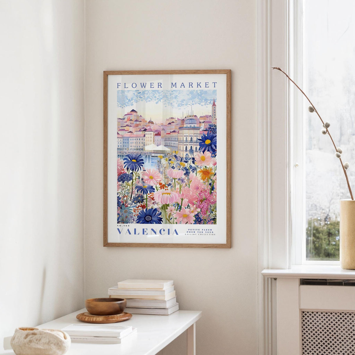 a framed poster of a colorful floral scene hanging on a wall, with a white table and a vase of flowers in the foreground.