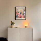 a white cabinet with a potted plant and a lamp on top, set against a wall with a framed poster of a flower market.