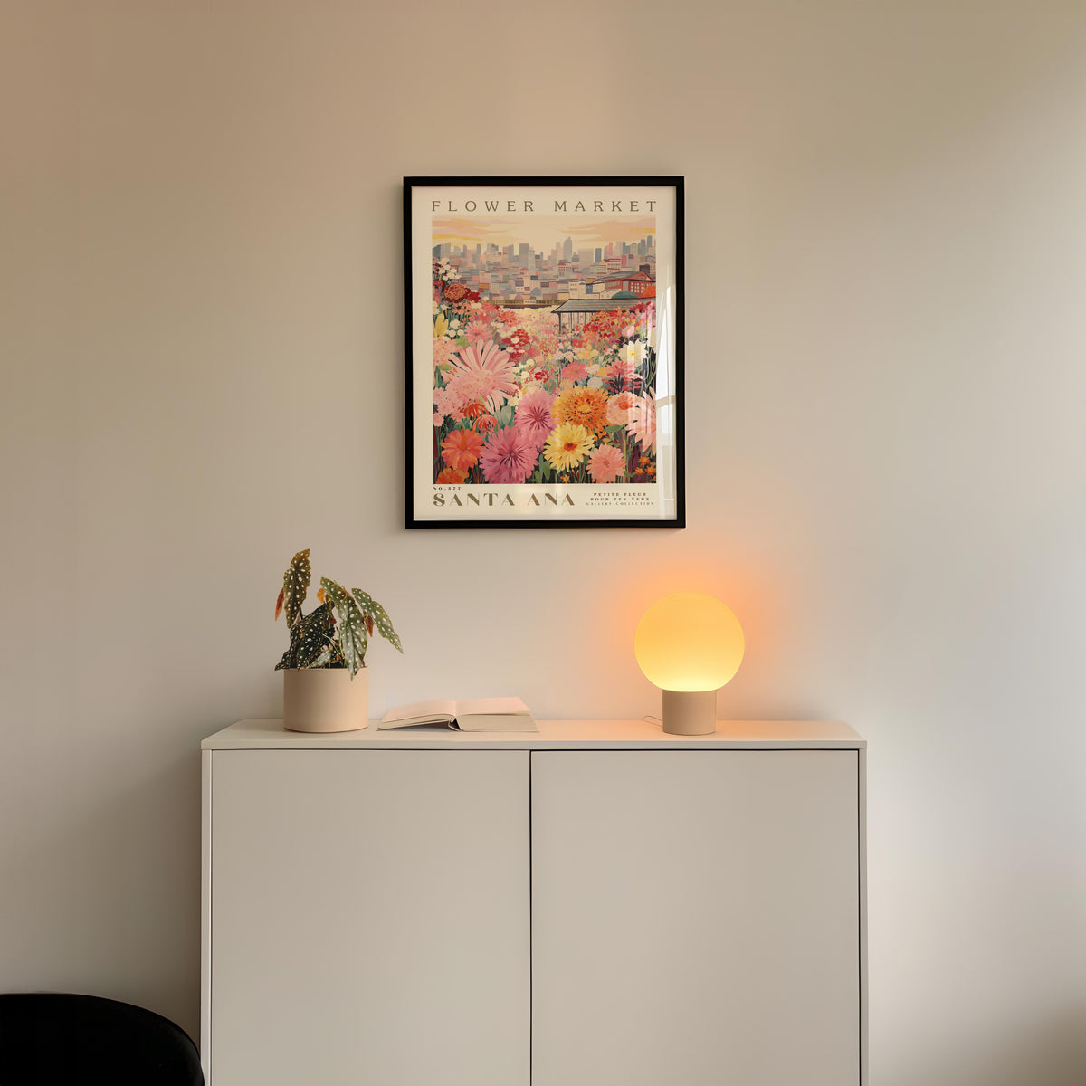 a white cabinet with a potted plant and a lamp on top, set against a wall with a framed poster of a flower market in Santa Ana, California.