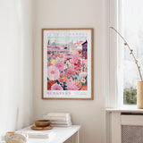 a framed poster of a colorful floral scene hanging on a wall, with a window and a small table with books and a vase in the foreground.