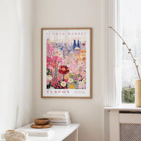 a framed poster of a colorful floral scene hanging on a wall, with a window and a small table with books and a vase in the foreground.
