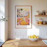 a kitchen scene with a framed poster of a colorful floral market hanging on the wall above a wooden counter. On the counter, there is a bowl of lemons and a plant. The kitchen has white tile backsplash and wooden shelves with various jars and containers.