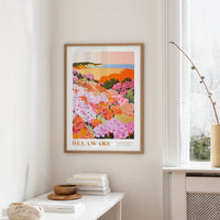 a framed poster of a colorful floral landscape hanging on a wall, with a white table and a vase of flowers in the foreground.