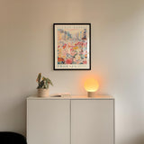 a white cabinet with a potted plant and a lamp on top, set against a wall with a framed poster of a cityscape above it.