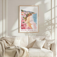a framed poster of Malibu Beach hanging on a wall above a white couch with a blanket draped over it.