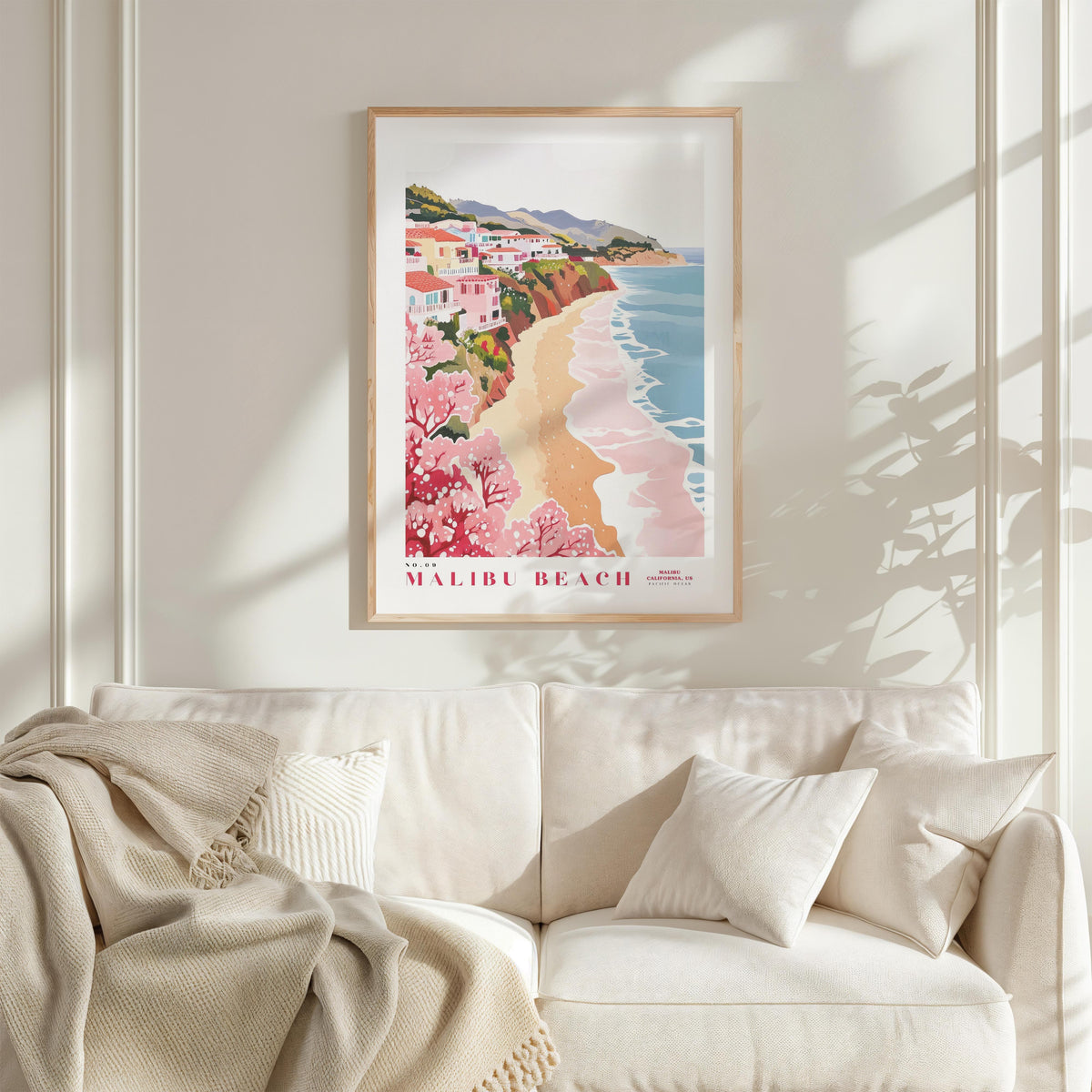 a framed poster of Malibu Beach hanging on a wall above a white couch with a blanket draped over it.