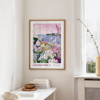 a framed poster of a colorful garden scene with flowers and a lighthouse in the background, hanging on a wall in a room with a window and a table with books and a vase.