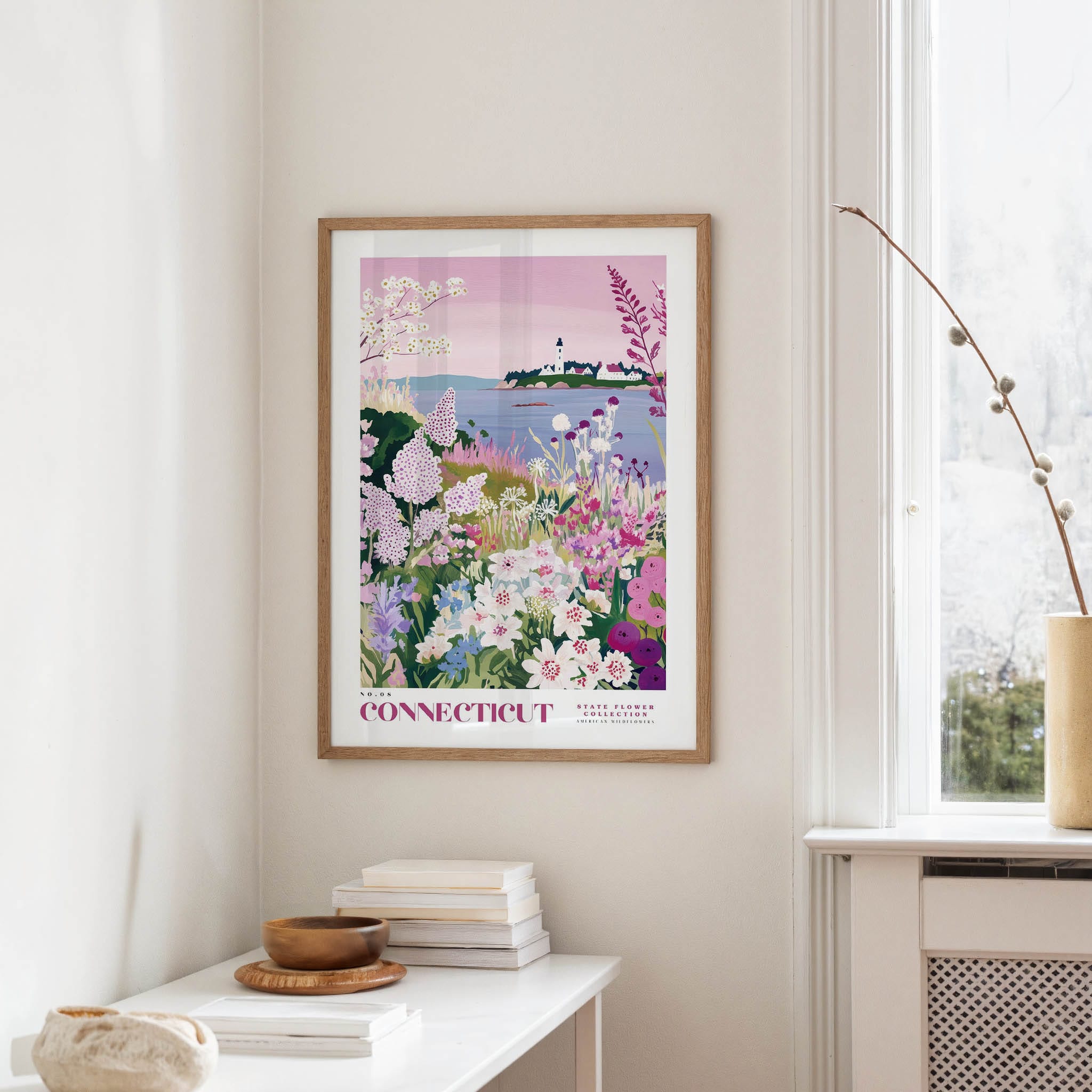 a framed poster of a colorful garden scene with flowers and a lighthouse in the background, hanging on a wall in a room with a window and a table with books and a vase.