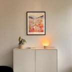 a white cabinet with a potted plant and a lamp on top, set against a wall with a framed poster of a floral scene hanging above it.