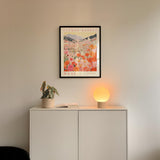 a white cabinet with a potted plant and a lamp on top, set against a wall with a framed poster of a floral scene hanging above it.