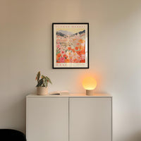 a white cabinet with a potted plant and a lamp on top, set against a wall with a framed poster of a floral scene hanging above it.