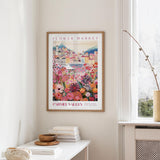 a framed poster of a colorful floral scene hanging on a wall, with a white table and a vase of flowers nearby.