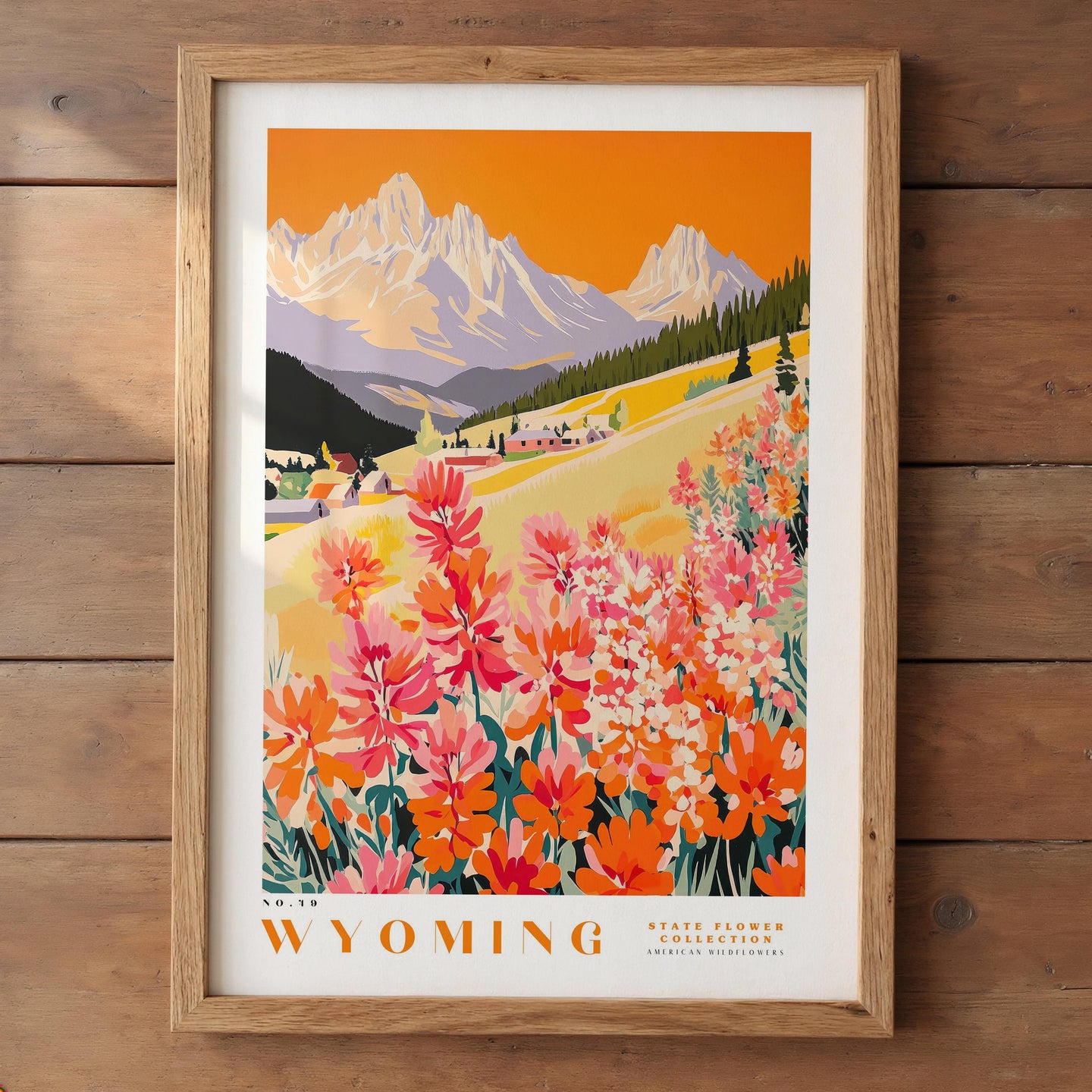 The image is a framed poster depicting a scenic landscape in Wyoming, featuring a mountain range, a village, and colorful wildflowers.