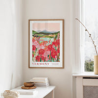 a framed poster of a vibrant floral landscape hanging on a wall, with a white table and a vase of flowers in the foreground.