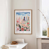 a framed poster of a cityscape with a floral design, hanging on a wall in a room with a white table and a window.