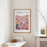 a framed poster of a colorful floral scene hanging on a wall, with a white table and a vase of flowers in the foreground.