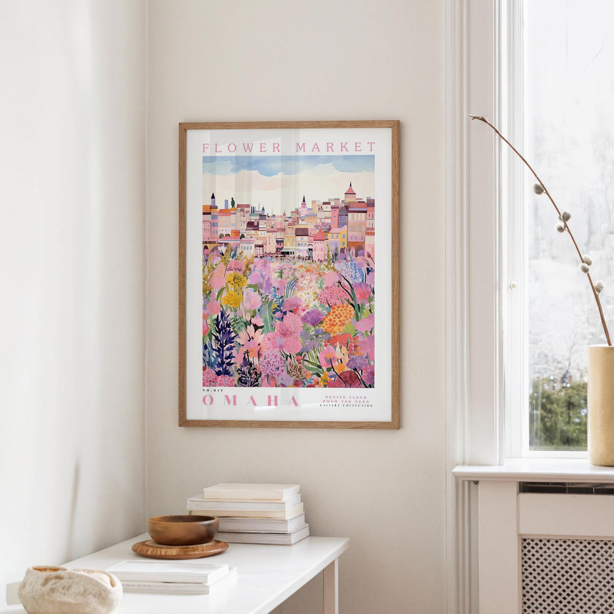 a framed poster of a colorful floral scene hanging on a wall, with a white table and a vase of flowers in the foreground.